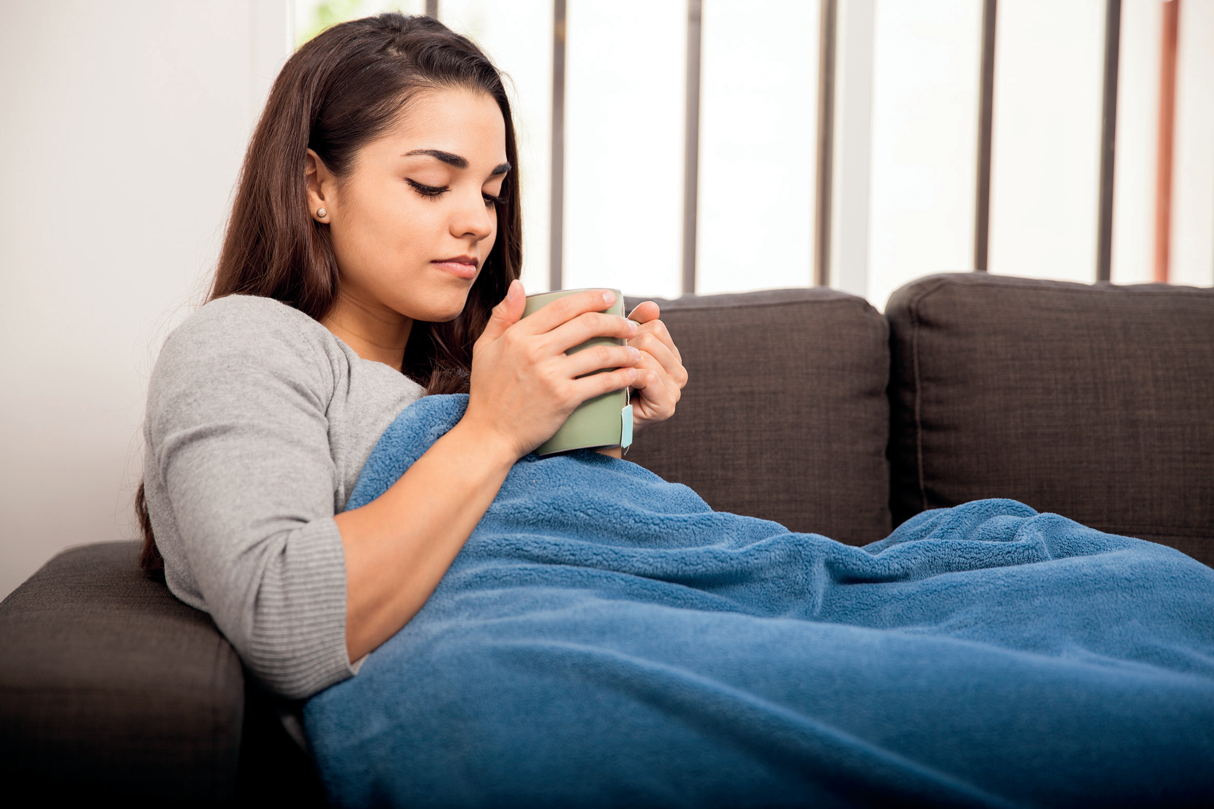 Junge Frau liegt auf dem Sofa mit Decke und einer Tasse Tee in den Händen