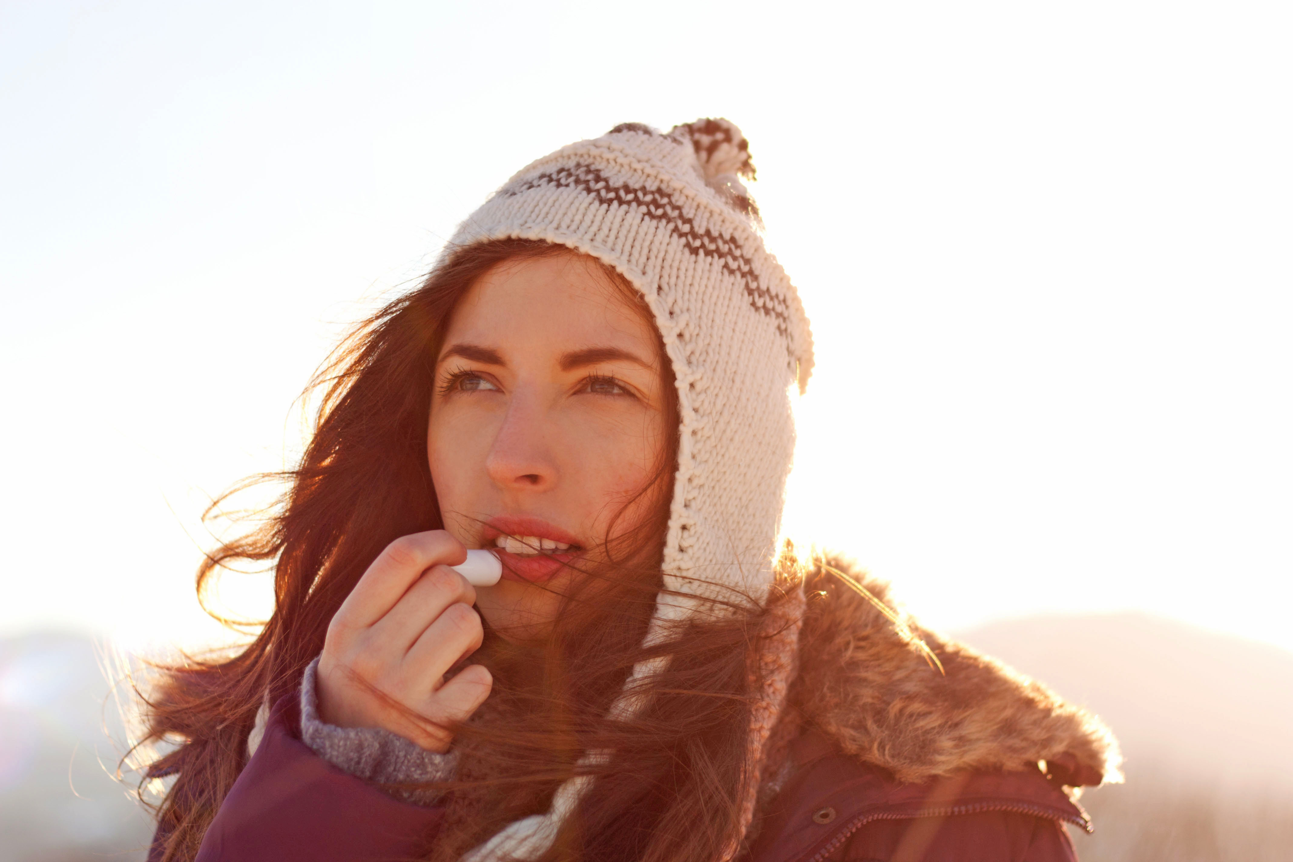 Junge Frau mit Mütze im Winter draußen cremt ihre Lippen ein